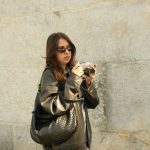 A fashionable young woman in sunglasses and leather jacket holding a camera outdoors. Urban setting with stone background.